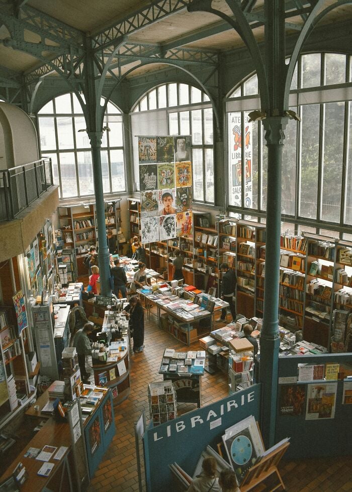 Biblioteca espa&ccedil;osa e grandiosa com grandes janelas arqueadas, altas colunas de metal e prateleiras cheias de livros e visitantes navegando.