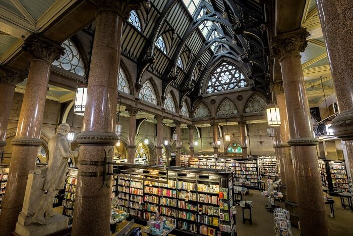 Interior grandioso de biblioteca com colunas imponentes, tetos em arco e prateleiras cheias de livros em um ambiente m&aacute;gico.