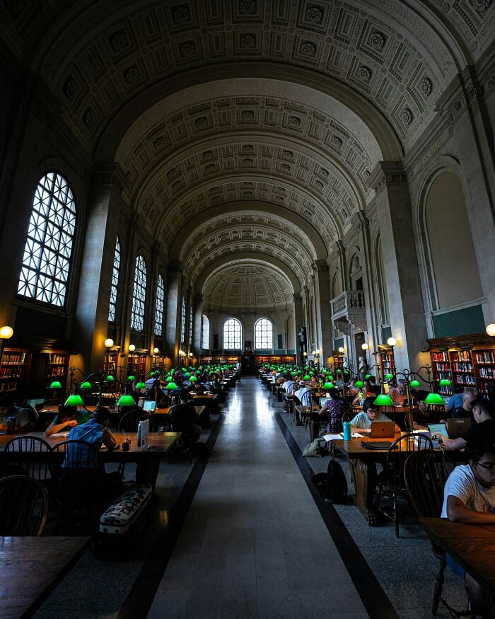 Interior grandioso de biblioteca com tetos altos em arco, grandes janelas e fileiras de mesas de estudo com lumin&aacute;rias verdes e leitores.