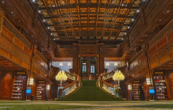 Interior grandioso de biblioteca com uma grande escadaria, paredes de madeira ornamentadas e estantes cheias de livros em um cen&aacute;rio m&aacute;gico.