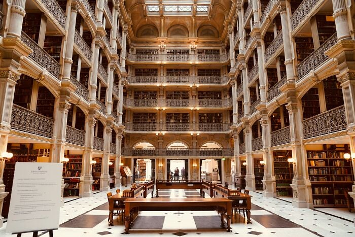 Interior grandioso de biblioteca com v&aacute;rios n&iacute;veis de estantes, colunas ornamentadas e uma ampla &aacute;rea de leitura com mesas e cadeiras de madeira.