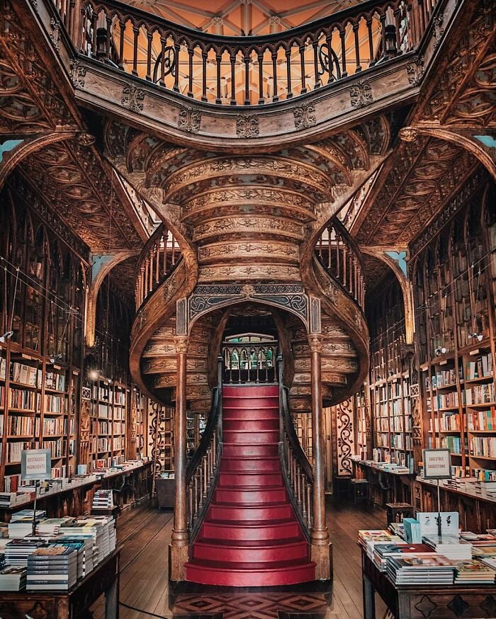 Biblioteca grandiosa e ornamentada com uma escadaria em espiral de madeira esculpida e fileiras de estantes cheias de livros ao redor.