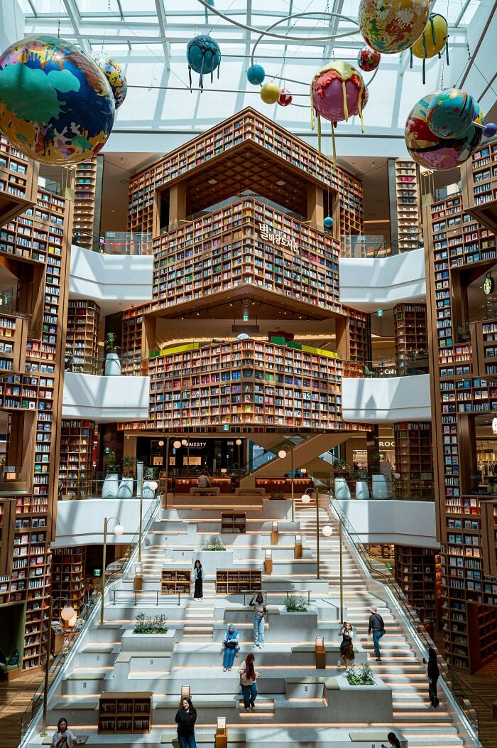 Interior grandioso de biblioteca com estantes imponentes, grande claraboia e esculturas coloridas de planetas penduradas no teto.